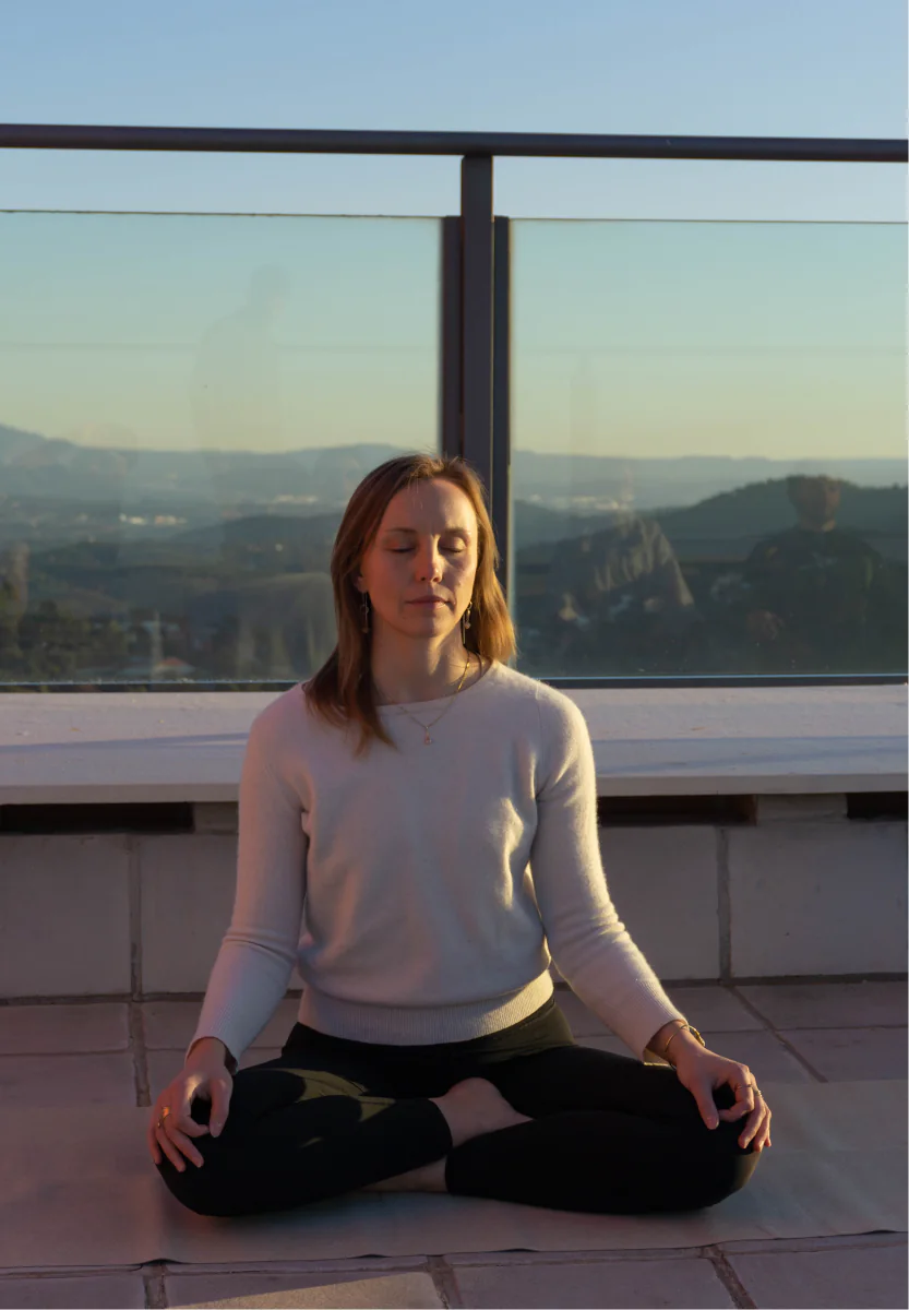 A woman meditates on the rooftop at sunset, with hills in the distance.