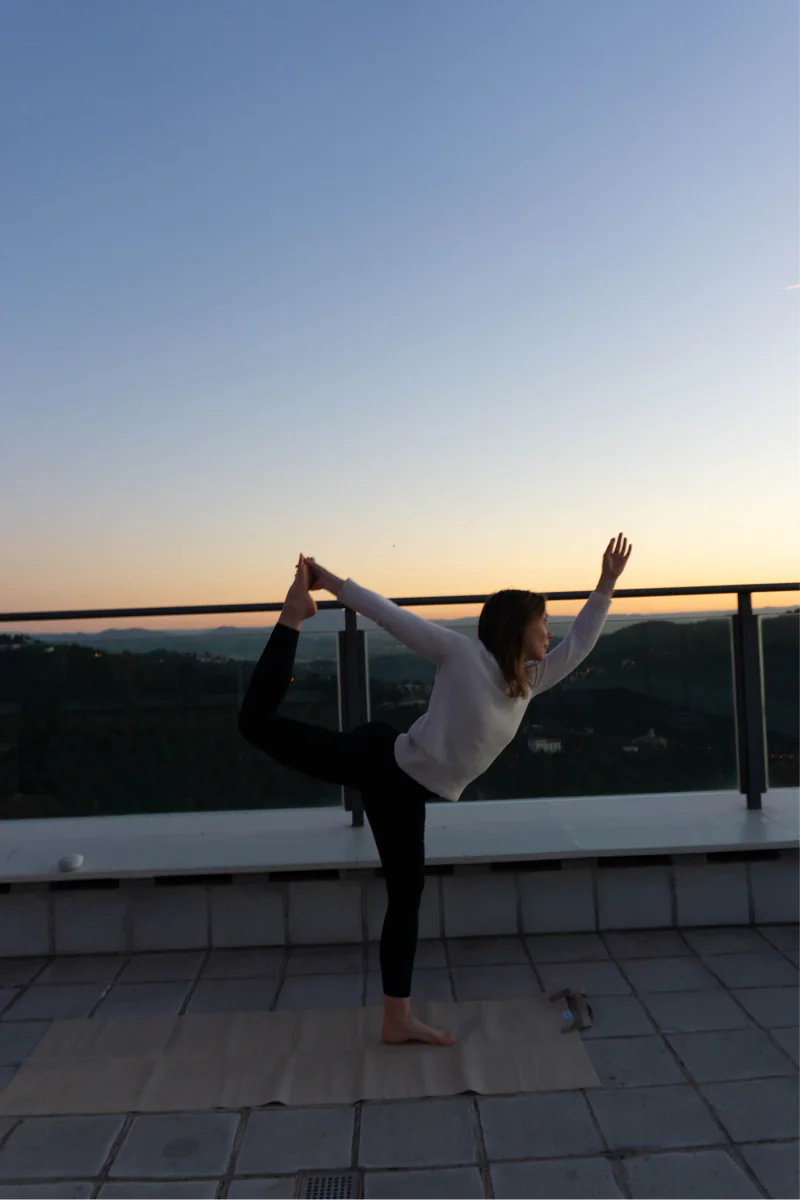 A woman holds a balancing yoga pose on the rooftop as the sun sets.